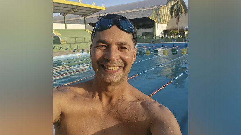 Homem sorrindo com óculos de natação na cabeça, em frente a uma piscina olímpica.