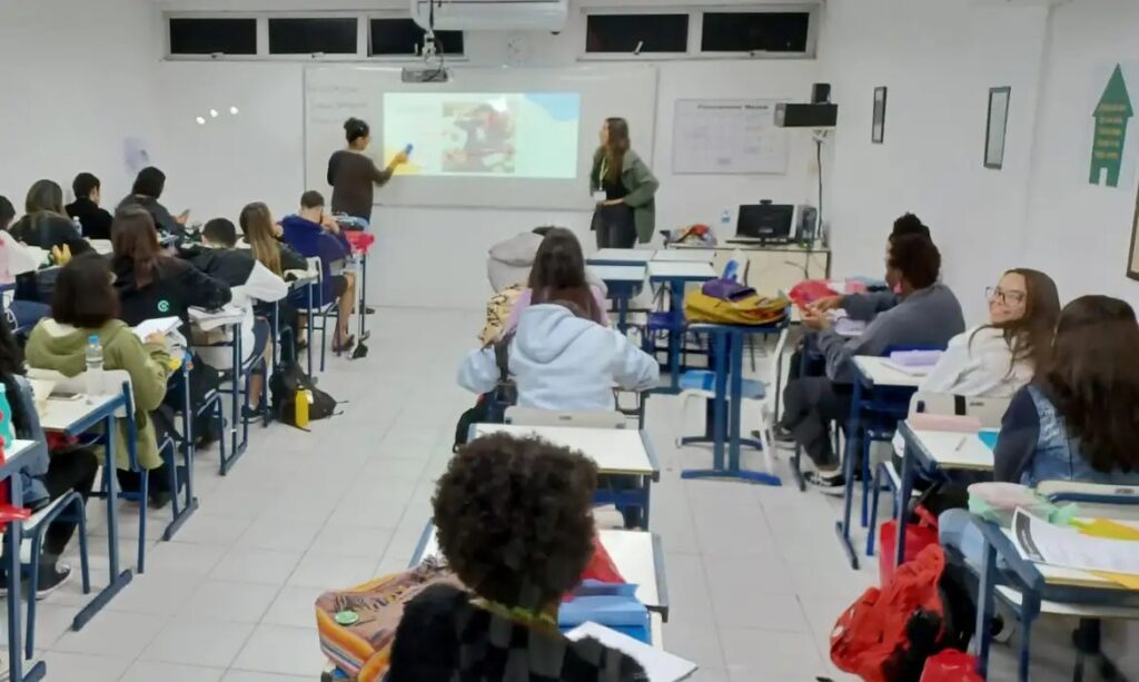 Sala de aula com duas professoras apresentando para alunos sentados em carteiras escolares.