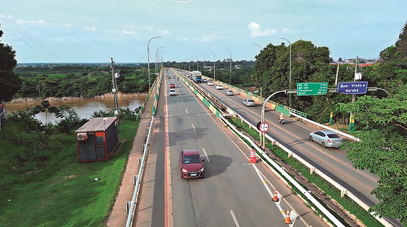 Ponte rodoviária movimentada sobre um rio barrento, cercada por vegetação densa.
