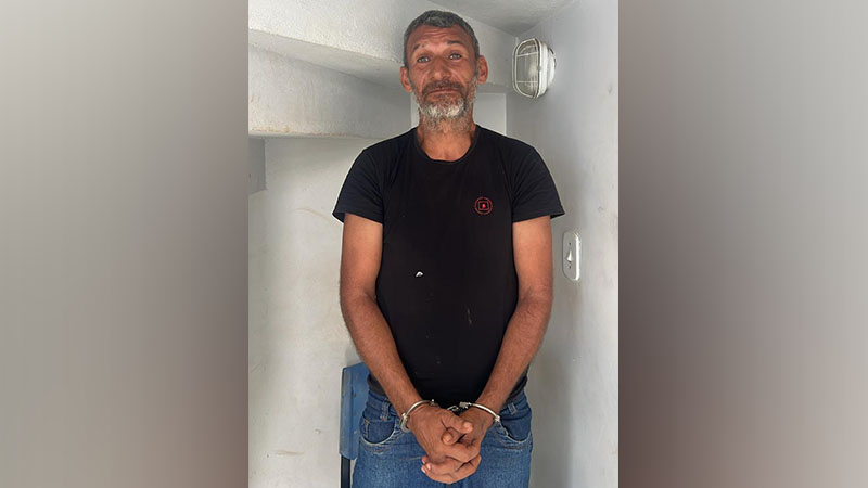 Homem de meia-idade, com barba grisalha e algemas nos pulsos, olhando para frente.