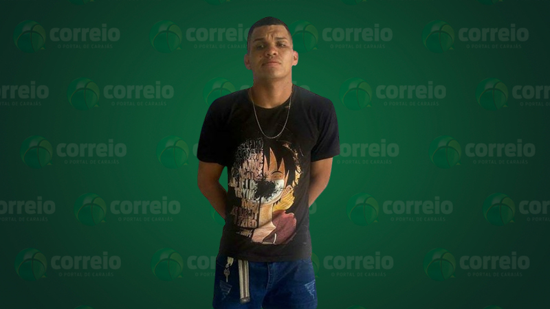 Homem em pé, de frente, com camiseta preta estampada, jeans e colar, em fundo verde com logo do "Correio".