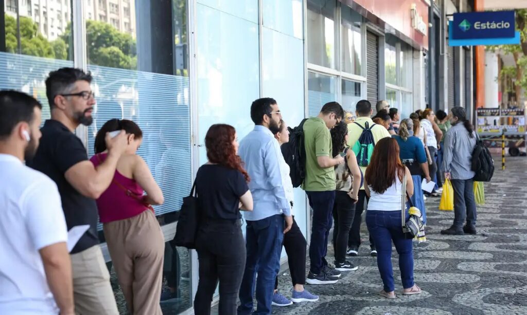 Fila de pessoas na calçada em frente a um prédio com janelas de vidro.