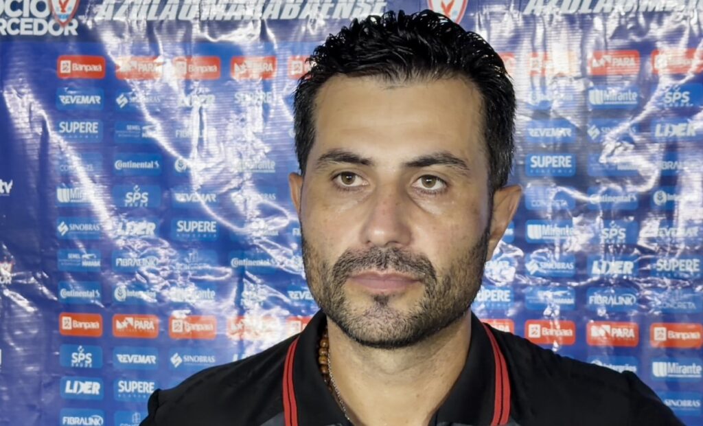 Homem com barba e cabelo escuro em frente a um banner de patrocinadores.