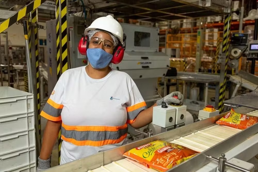 Mulher com EPI completo (capacete, máscara, óculos, protetor auricular) operando máquina em fábrica, com um pacote de caldo na esteira.