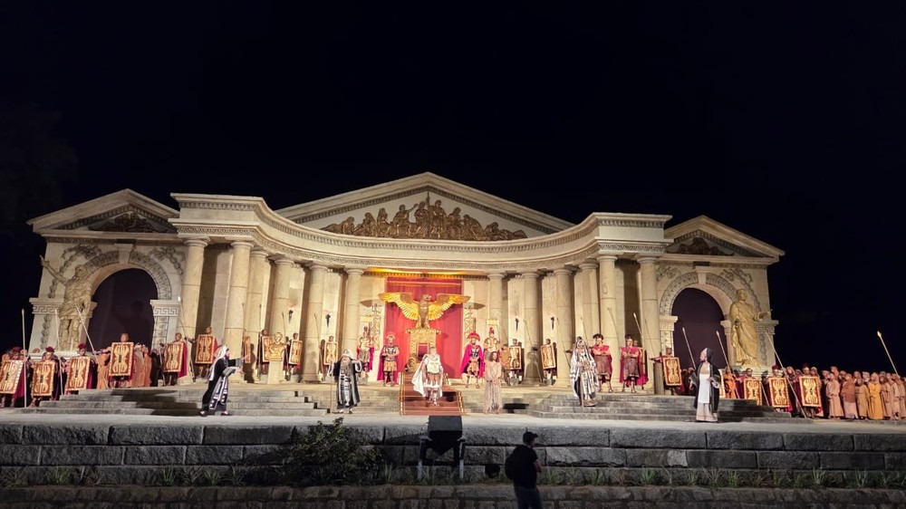 Uma encenação noturna de uma peça teatral histórica com atores em trajes romanos em um palco grandioso.