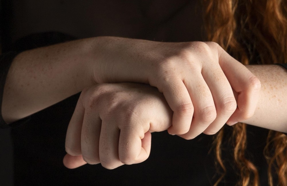 Close-up de duas mãos de pele clara, uma formando um punho e a outra apoiada sobre ela, com fundo escuro e cabelo ruivo ao fundo.