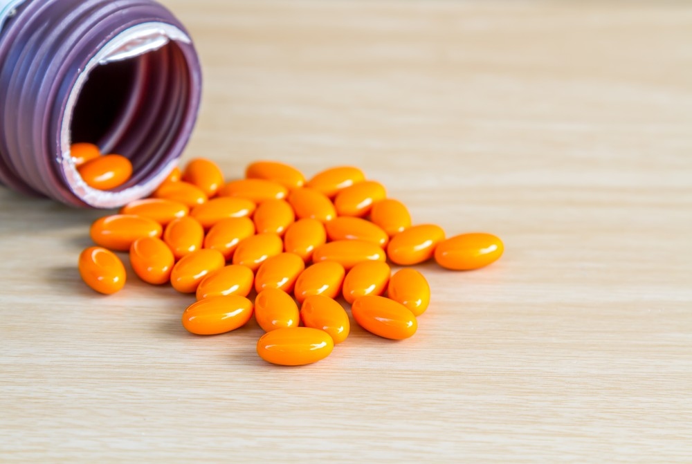 Pílulas ovais alaranjadas derramadas de um frasco roxo sobre uma mesa de madeira clara.