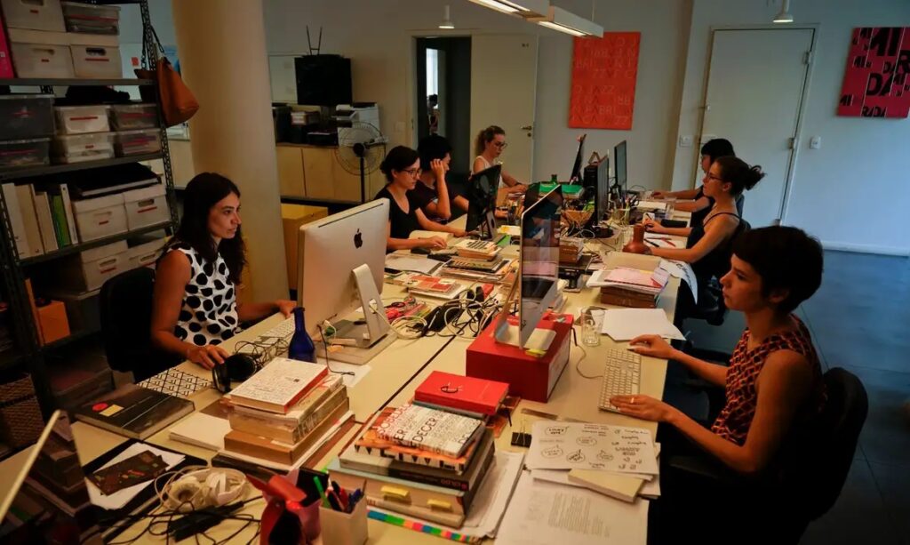 Mulheres trabalhando em um escritório movimentado com computadores e pilhas de livros.