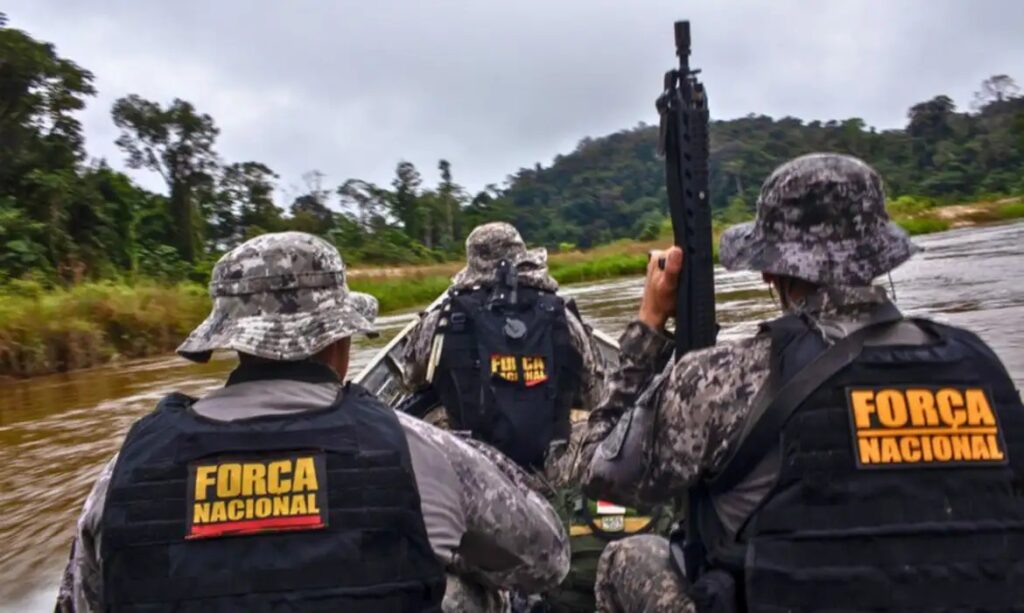 Membros da Força Nacional em barco com fuzil, patrulhando um rio na floresta.