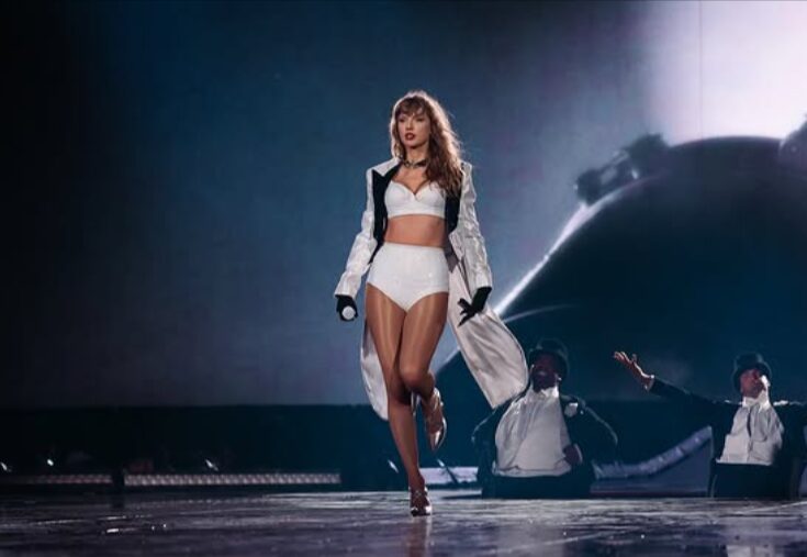 Taylor Swift no palco, usando um conjunto branco com casaco longo e segurando um microfone.