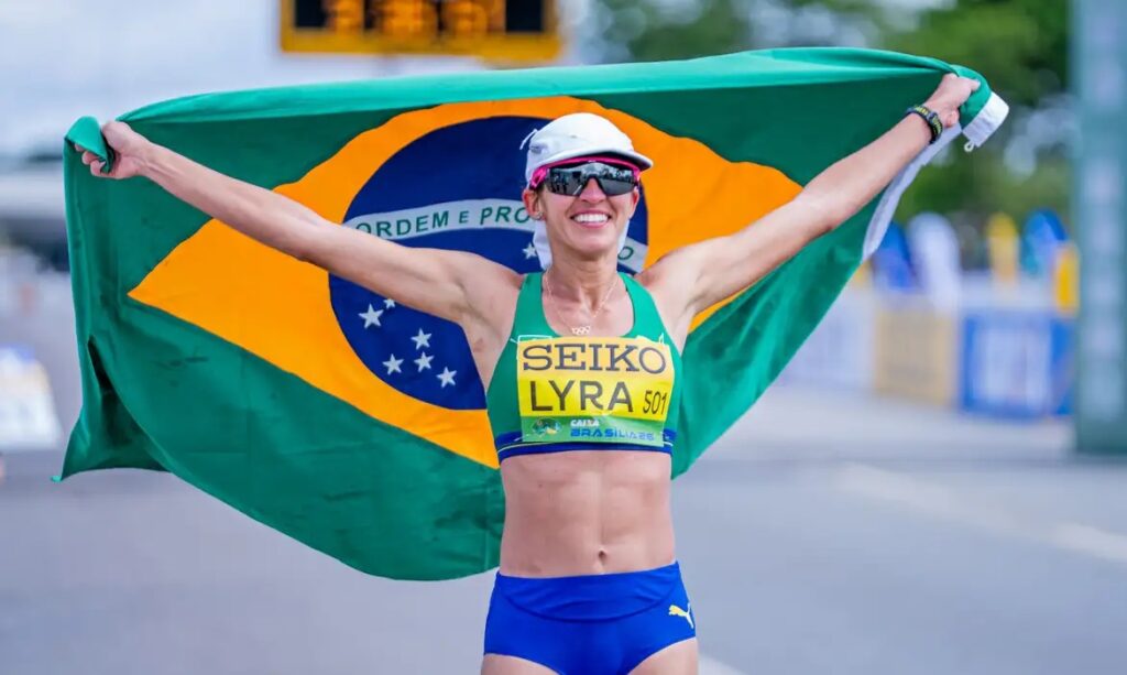 Atleta feminina sorrindo e segurando a bandeira do Brasil.