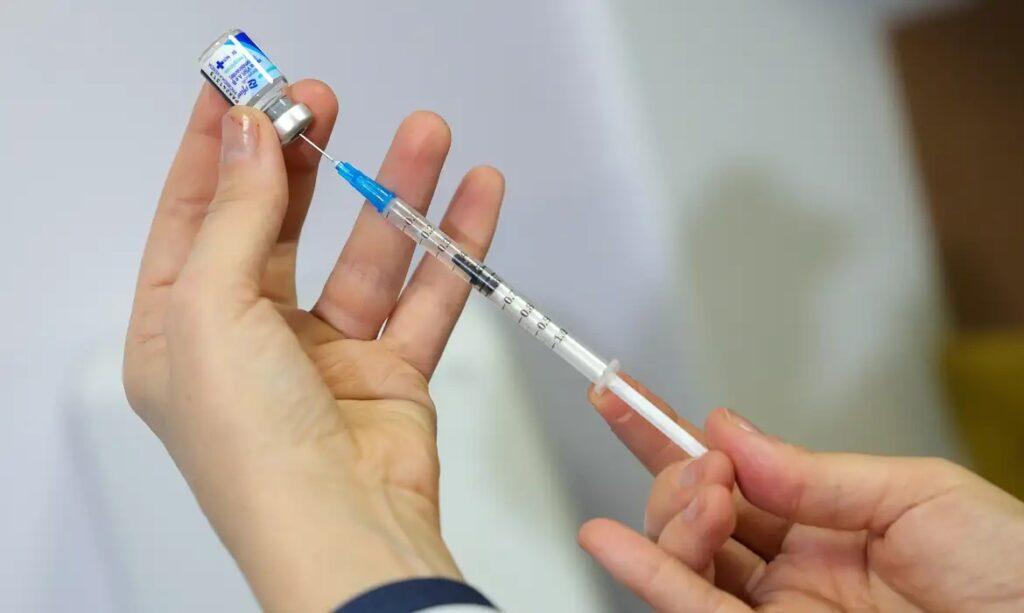 Mãos segurando seringa e frasco de medicamento azul, com a agulha inserida no frasco.