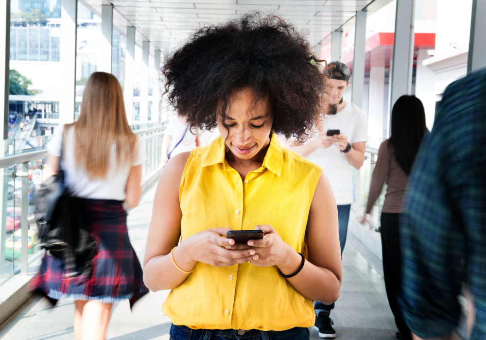 Mulher jovem com cabelo afro encarando o celular enquanto caminha por uma passarela.