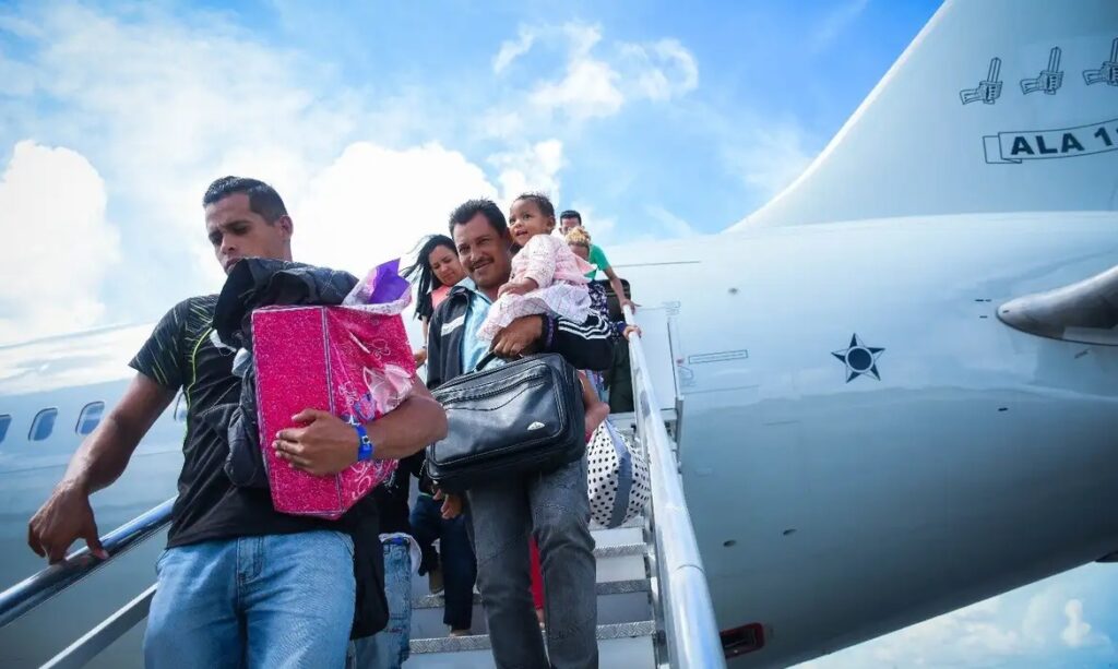 Homem sorrindo segurando criança no colo e bolsa, descendo escada de avião, com outra pessoa carregando uma caixa rosa ao lado.