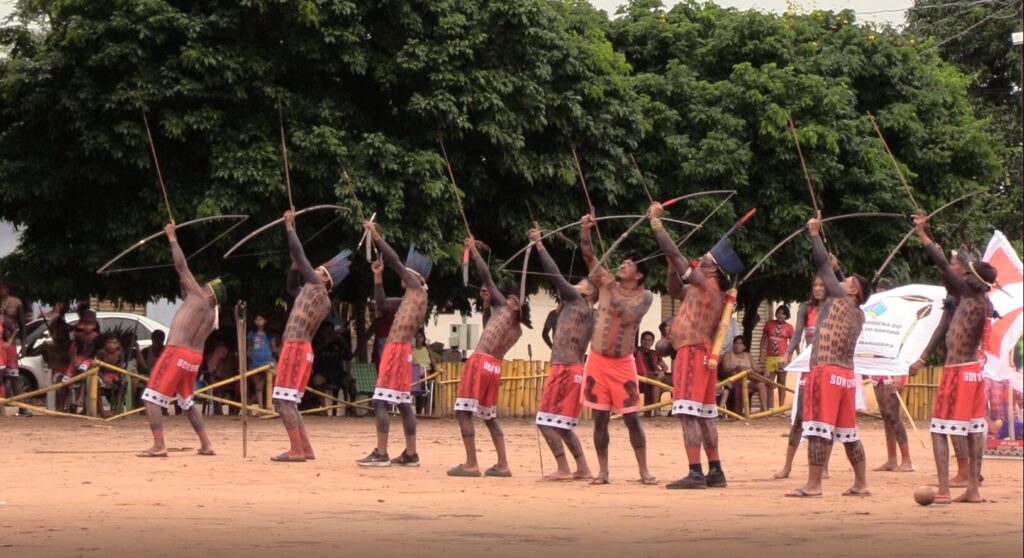 Sete homens indígenas pintados com arcos e flechas apontados para o céu, em evento cultural.
