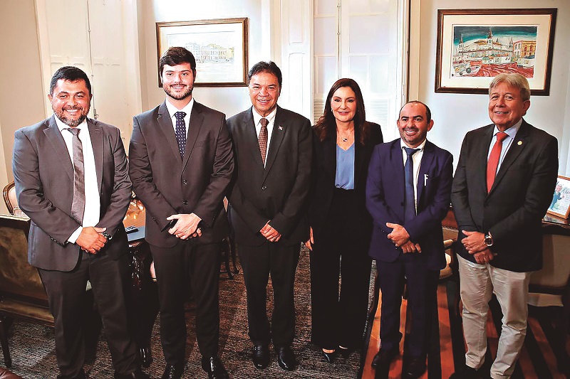 Grupo de seis profissionais, cinco homens e uma mulher, em trajes formais, sorrindo em um ambiente de escritório.
