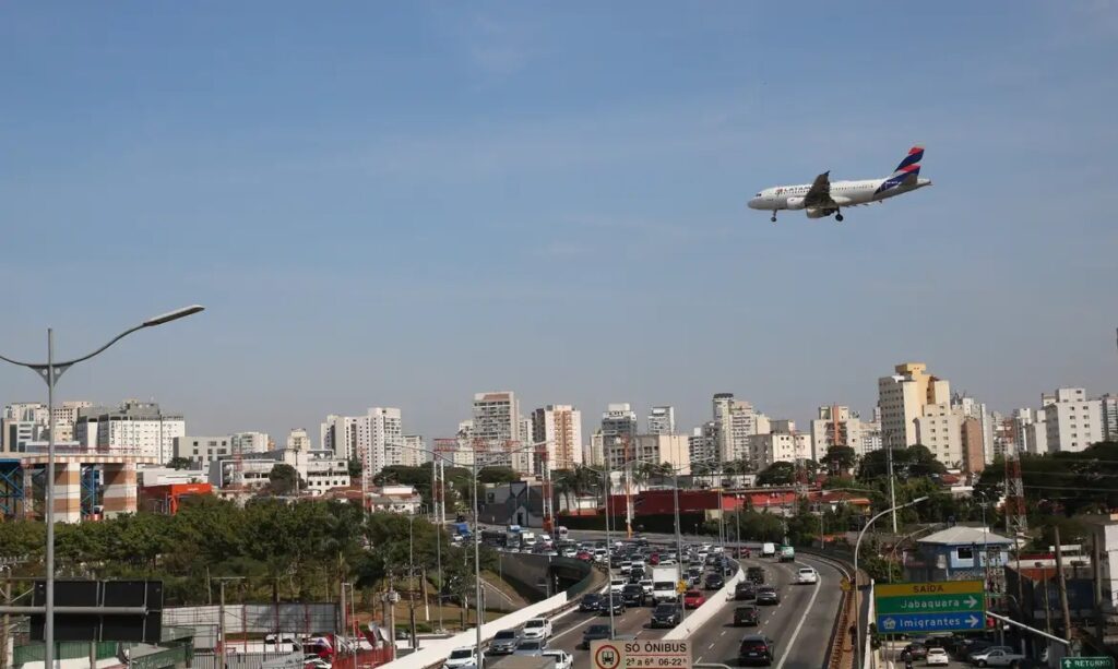 Avião da LATAM sobrevoando uma rodovia movimentada com tráfego intenso e prédios de cidade ao fundo sob céu azul.