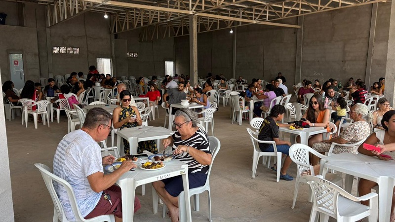 Pessoas de diferentes idades comendo em mesas e cadeiras de plástico em um grande salão.