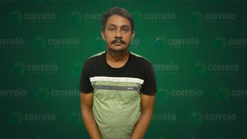 Homem de meia-idade com barba e bigode, usando camiseta verde e preta, em frente a um fundo com o logotipo "correio".