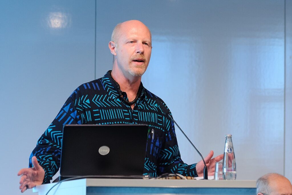 Homem calvo com camisa estampada azul e preta falando em um pódio com laptop e microfone.