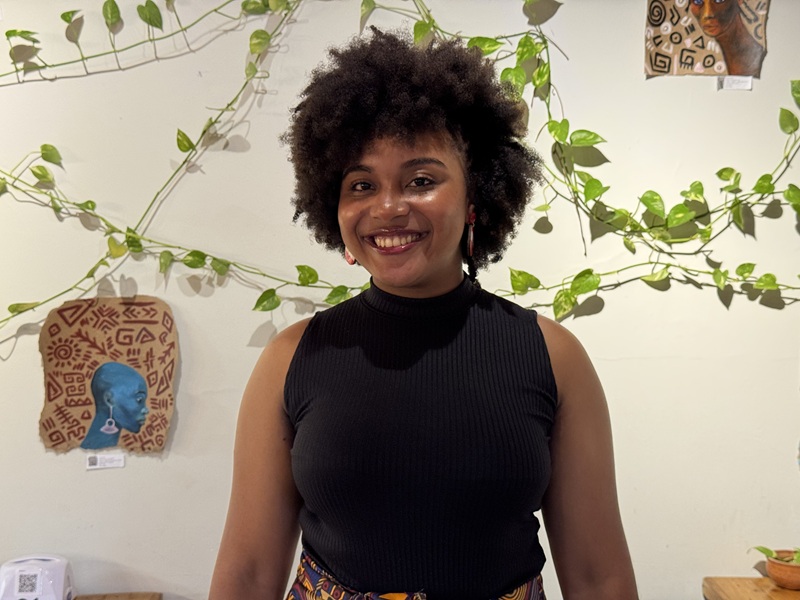 Jovem mulher negra sorrindo com cabelo afro, em frente a parede com plantas e arte africana.