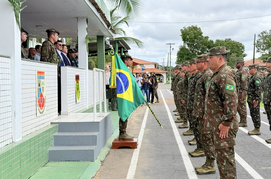Militar segurando a bandeira do Brasil em uma formatura, com outros soldados e autoridades observando.