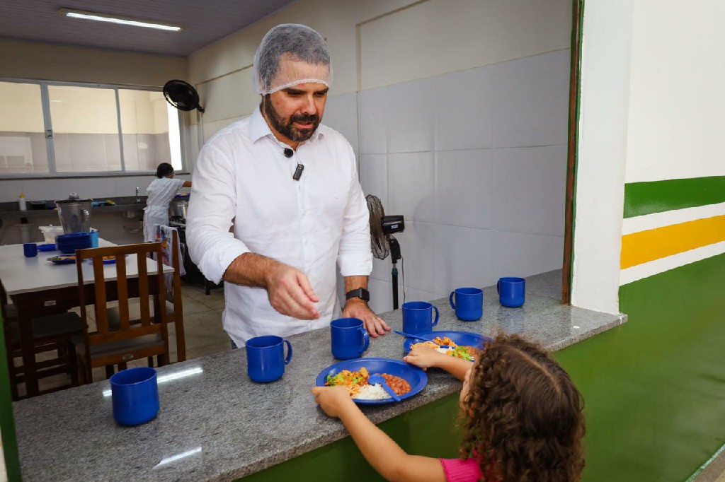 Homem de touca serve refeição balanceada para criança em refeitório escolar.