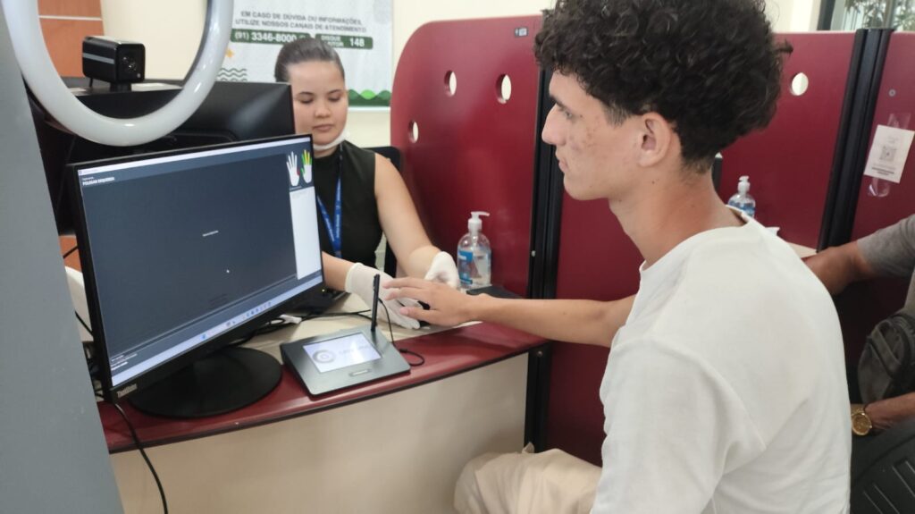 Homem fazendo coleta de biometria digital em balcão de atendimento, auxiliado por uma atendente.