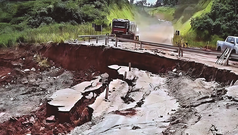 Trecho de estrada com asfalto colapsado e grande erosão, com um caminhão ao fundo.