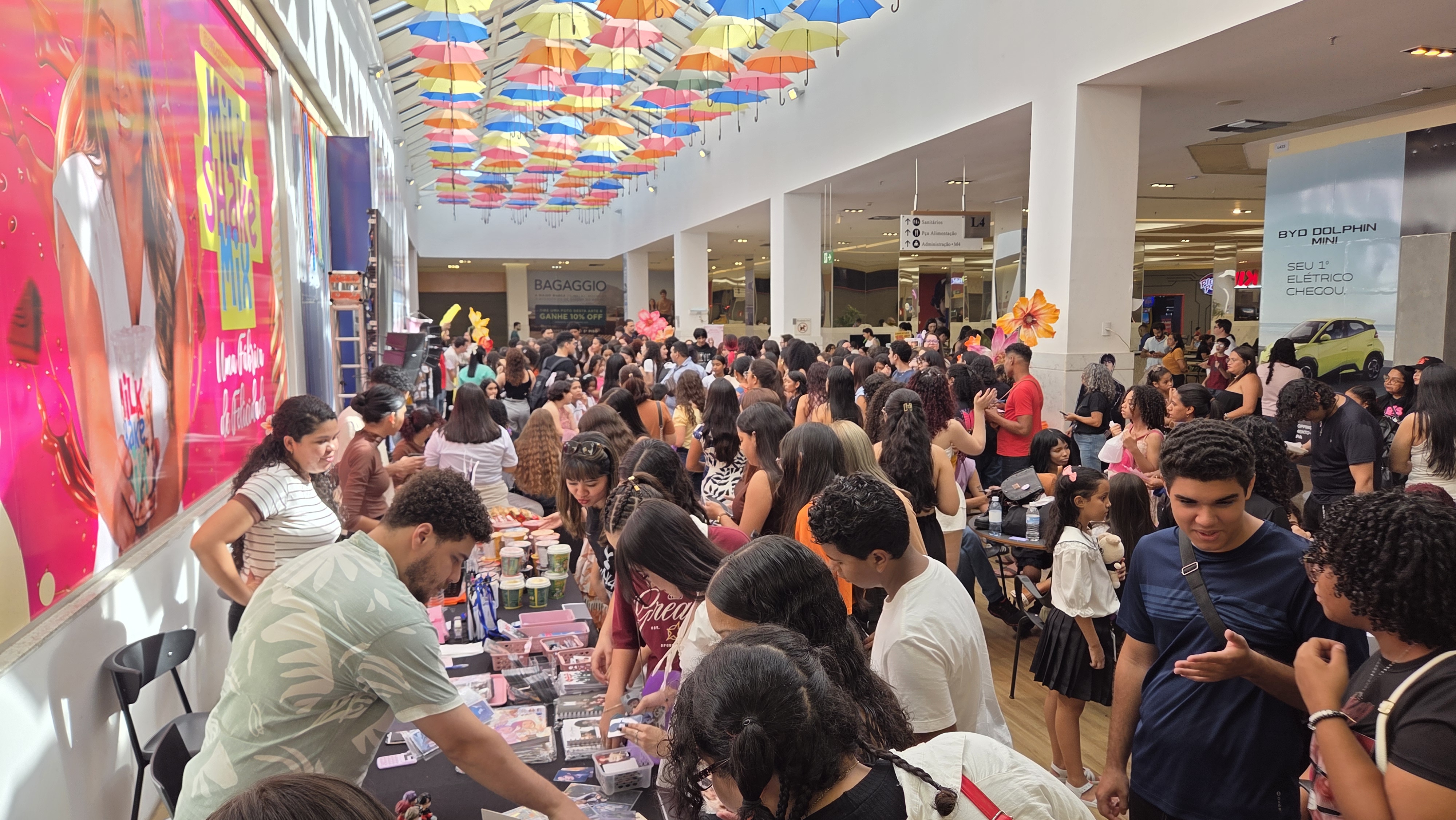 Multidão em um evento ou feira em um shopping, com guarda-chuvas coloridos pendurados no teto.