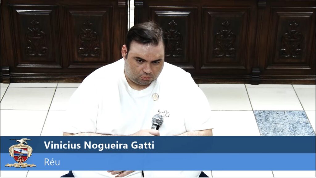 Homem de camiseta branca, Vinícius Nogueira Gatti, segurando um microfone e olhando para baixo com expressão séria.