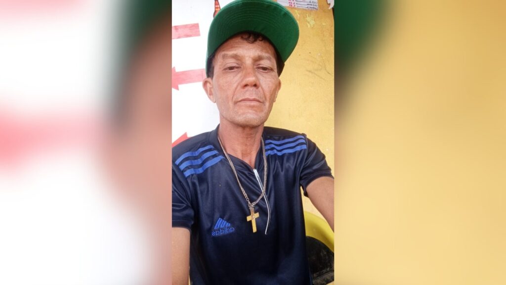 Homem de boné verde e camiseta azul com um colar de cruz dourada, olhando para a câmera.