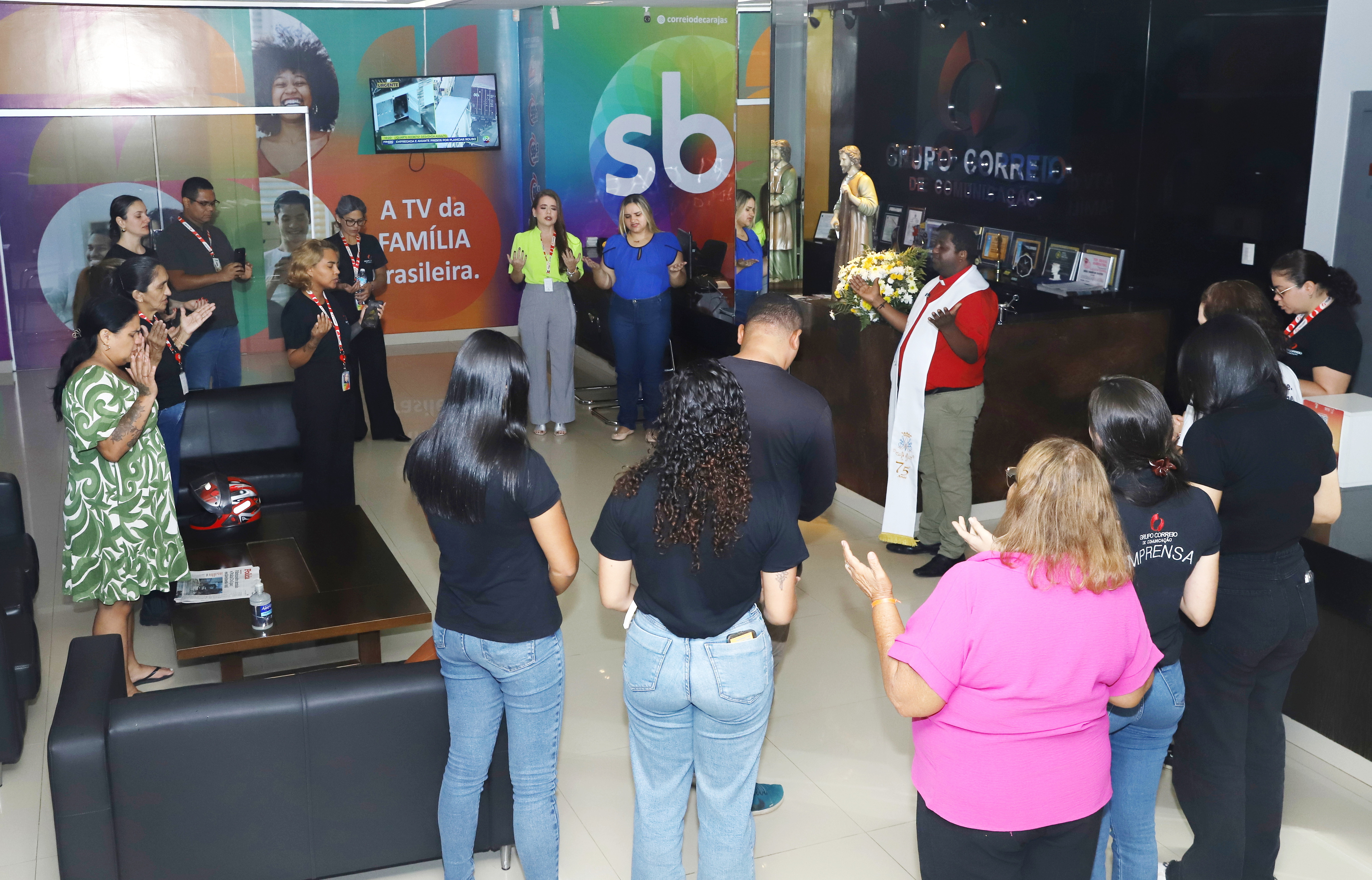 Grupo de pessoas em um ambiente de escritório com logotipos do SBT e Grupo Correio, sendo lideradas em um momento de oração por um homem vestindo batina.