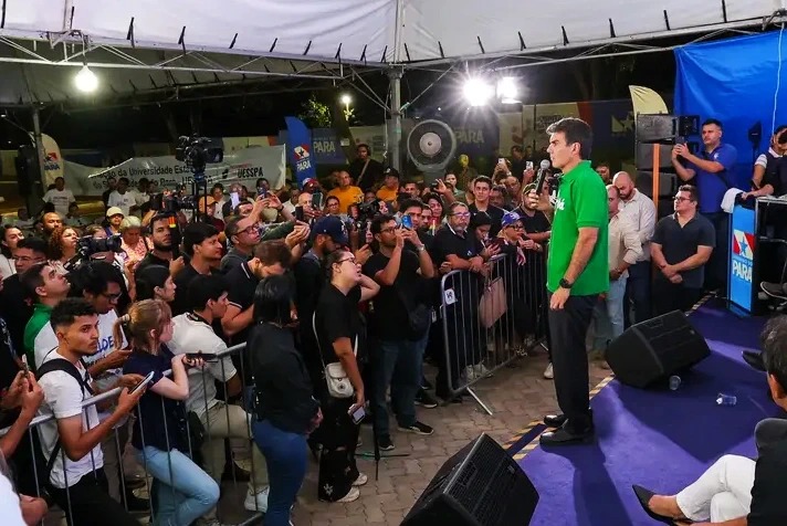 Homem de camisa verde discursando em palco para uma multidão, com muitas pessoas filmando com celulares.