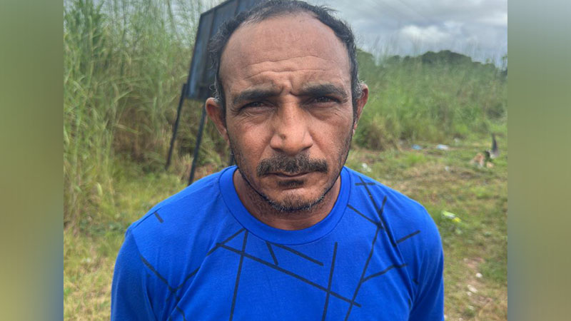 Homem com camisa azul e bigode olhando para a câmera em área externa.