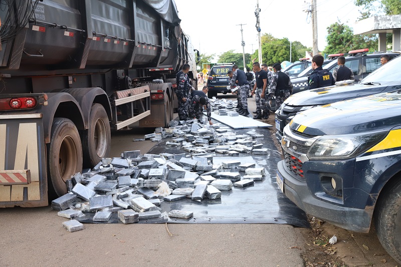 Apreensão de grande quantidade de drogas pela PRF, com pacotes espalhados no chão ao lado de um caminhão e viaturas policiais.