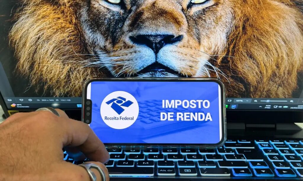Smartphone com app da Receita Federal e Imposto de Renda sobre teclado de notebook, com rosto de leão ao fundo.