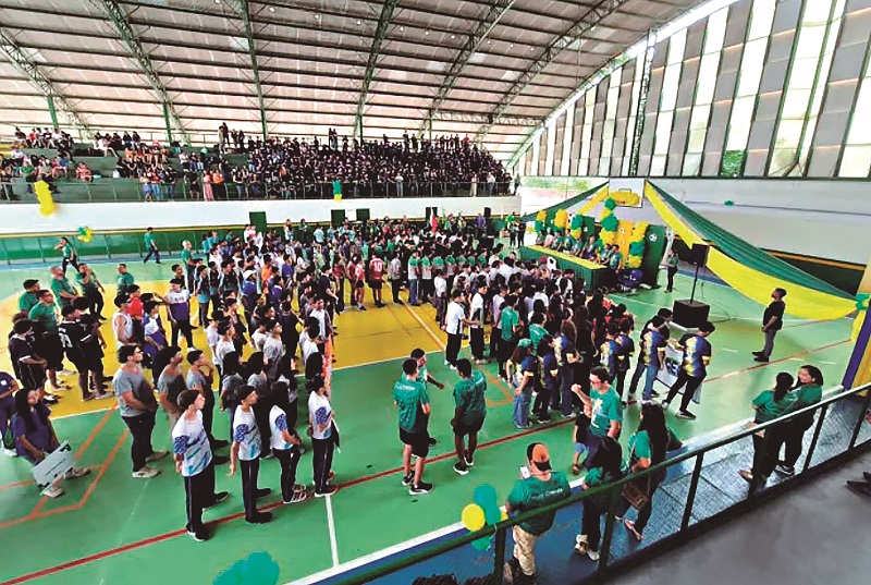 Vista aérea de um ginásio lotado com jovens no centro da quadra e arquibancadas cheias.