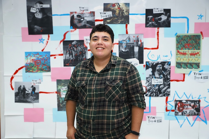 Pessoa sorridente de camisa xadrez em frente a uma parede decorada com arte contemporânea e referências visuais.