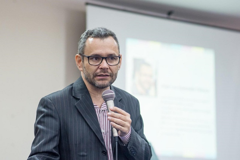 Homem de óculos e paletó listrado falando ao microfone durante uma apresentação.