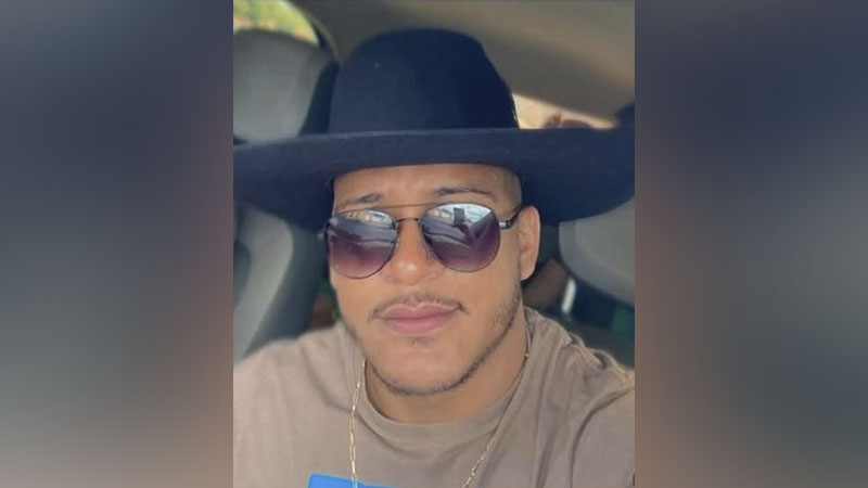 Homem de pele clara, chapéu fedora preto, óculos de sol aviador e corrente dourada no pescoço, dentro de um carro.