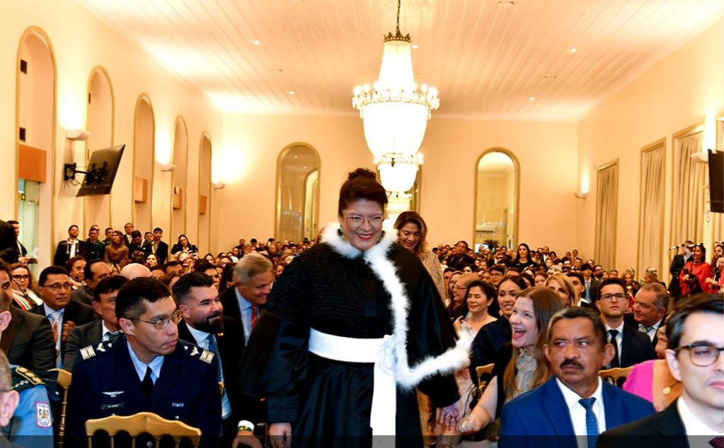 Mulher sorridente em beca preta com estola branca, caminhando em evento formal.