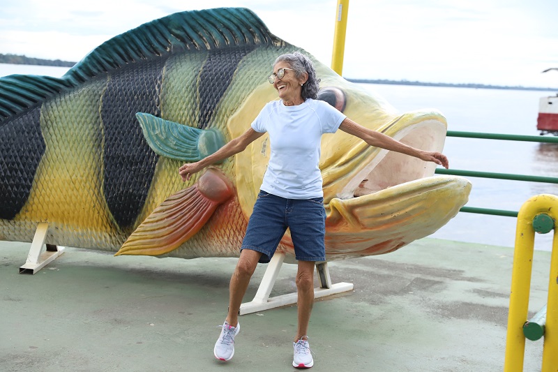 Mulher idosa sorrindo e posando alegremente ao lado de uma estátua gigante de peixe, com um rio ao fundo.