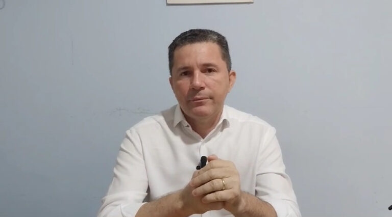 Homem de camisa branca com microfone de lapela, mãos unidas, olhando para frente.