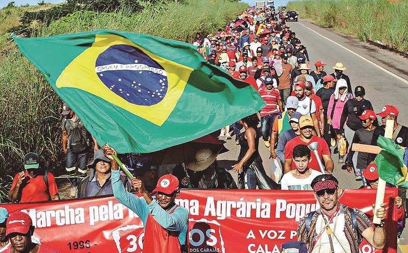 Multidão marcha com bandeira do Brasil e faixa pela reforma agrária.