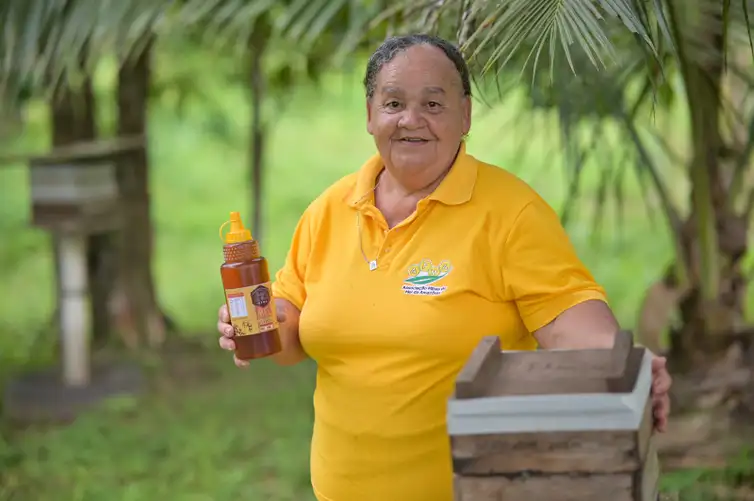 Mulher sorridente segurando uma garrafa de mel e uma parte de colmeia em ambiente natural.