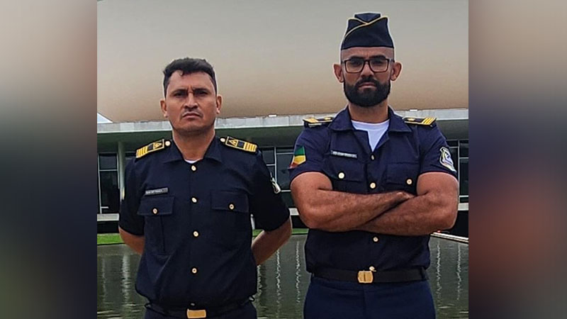 Dois homens em uniforme azul-marinho posam em frente a um edifício moderno e um espelho d'água.