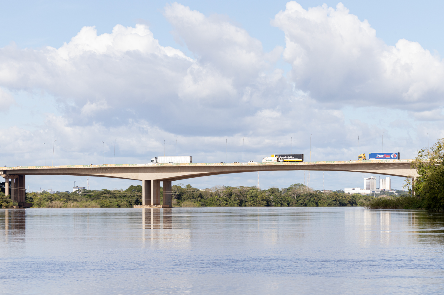 Ponte sobre rio com caminhões e carros sob céu nublado.