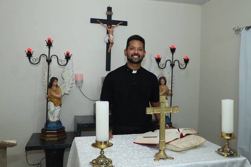 Sacerdote sorridente em altar doméstico com crucifixos e estátuas de anjos.