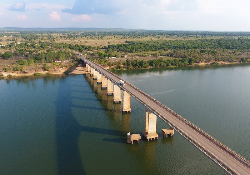 Ponte rodoviária sobre um rio extenso com um ônibus passando, em meio a paisagem rural e vegetação.